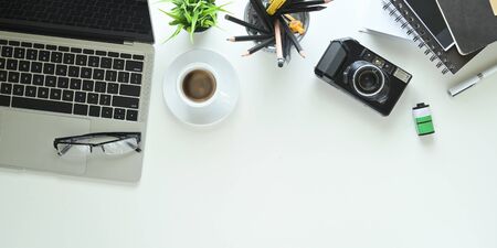 Top view image of coffee cup putting at the center of white working desk that surrounded by computer laptop, eye glasses, potted plant, coffee cup, pencil holder, camera, film and stack of notebook.の写真素材