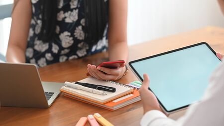 Cropped image of businessman holding a white blank screen computer tablet while sitting in front of casual woman that sitting and holding mobile in hand at the wooden working desk.の写真素材
