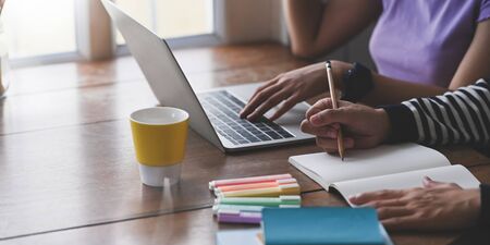 Cropped image of young couple doing their homework together while sitting in front of computer laptop that putting on wooden working desk and surrounded by coffee cup, marker pens and notebook.の写真素材