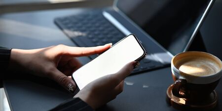 Cropped image of hands holding a cropped black smartphone with white blank screen over working desk that surrounded by office equipment as background.の写真素材