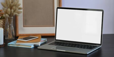 Photo of white blank screen computer laptop putting on black working desk and surrounded by stack of...
