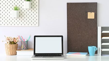 A white blank screen computer laptop is putting on a white workspace surrounded by office equipment.