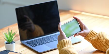A woman's hands using a smartphone while sitting in front of a computer laptop.