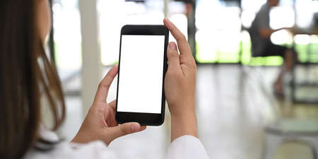 Behind of woman is holding a white blank screen smartphone while standing over blurred background.