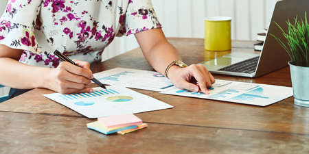 Cropped image of a woman is working with graphic charts while sitting at the white working desk.