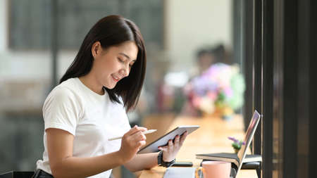 A beautiful woman is sketching on a computer tablet in hand while sitting in a comfortable coffee cafe.の写真素材