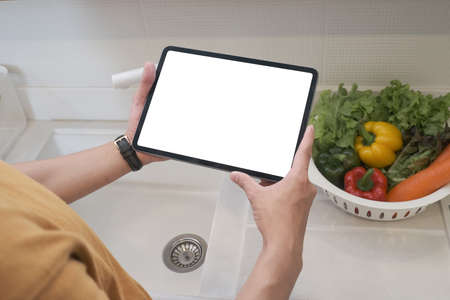Woman holding a tablet in kitchen with vegetable at the side.