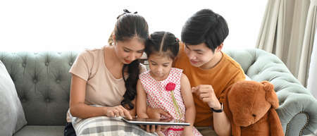 Cheerful kids and their parents in casualwear relaxing on couch in living room and using digital...