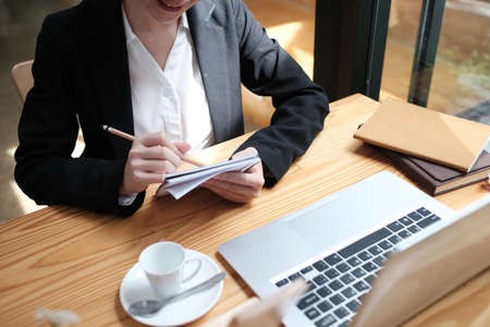 Young business woman sitting at table and taking notes in notebook on wooden desk.の写真素材