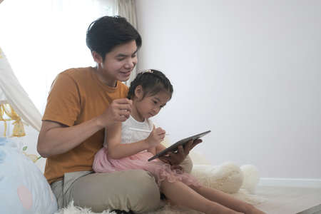 Father and daughter using digital devices on bed in bedroom.