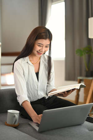 A happy woman reading a book while using laptop on sofa in her living room.