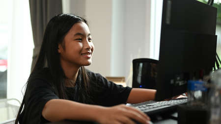 A smiling little school girl playing online game on computer or using educational applications on electronic device or watching cartoons at home.の写真素材