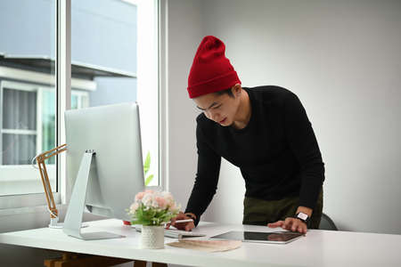 A young male designer is examining paper with color samples while working on project in modern...