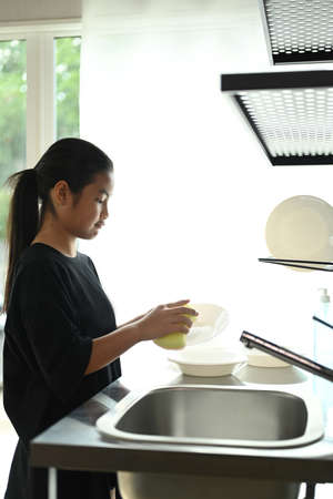 A cute teenage girl is washing dishes in the kitchen at home.