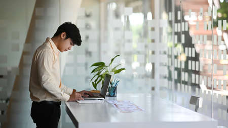 A businessman is working online with a laptop and writing down notes while sitting in a modern...