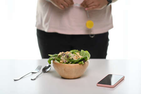 A healthy bowl of salad on white table with a woman measuring her waist size with tape measure.の写真素材