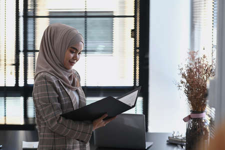 Portrait of muslim businesswoman in hijab is holding document and smiling to camera while standing by the window.の写真素材
