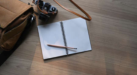 Top view of photographer or artist workspace with notebook, pencil, camera and bag on wooden table.