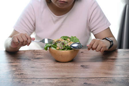 Cropped shot of overweight woman is tired of diet restrictions eating green salad sitting at table.の写真素材