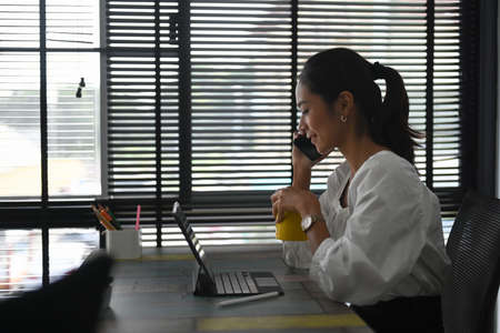 Happy cheerful young woman making answering call by cellphone while sitting in her office room.