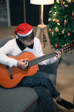 Portrait of young woman in Santa hat playing guitar on sofa at home during coronavirus quarantine long distance celebration.の写真素材
