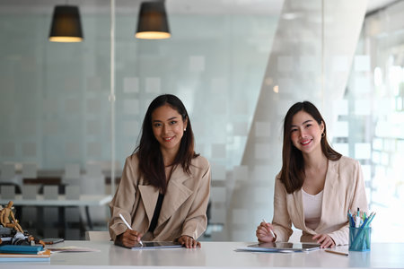Two cheerful businesswoman colleagues working together on new project and smiling to camera in modern office.の写真素材