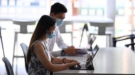 Side view of businesspeople wearing protective mask working with tablet computer and sitting...