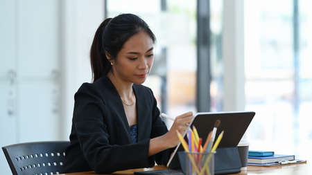 Beautiful businesswoman holding stylus pen writing information on tablet computer in modern office.