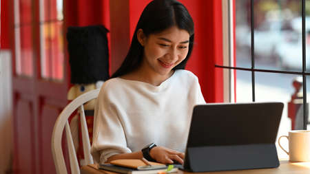 Smiling young businesswoman sitting near window in coffee shop and working on computer laptop.