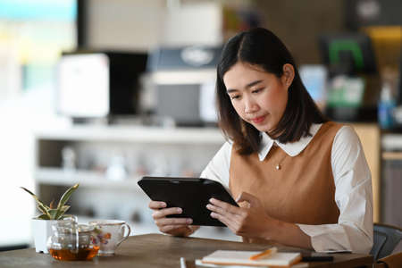 Young female holding digital tablet and thinking about her project while sitting in modern coffee...