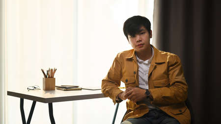 Young asian man in yellow jacket looking at camera and sitting near window at his home office.