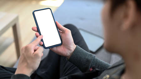 Close up view of young man holding mobile phone with empty screen while sitting on sofa at home.