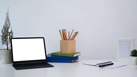 Stylish workplace with computer tablet, plant, notebook, pencils holder and book on white desk.