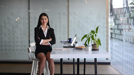 Confident businesswoman sin black suit sitting with arms crossed in modern office.の写真素材