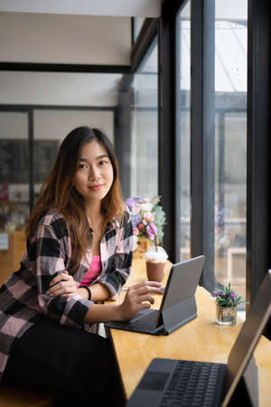 Portrait of young designer sitting near window in coffee shop with her computer table and smiling to camera.の写真素材