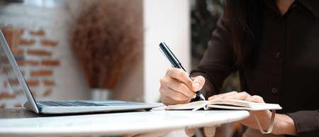 Cropped shot of young female freelancer working with laptop computer and writing notes into...
