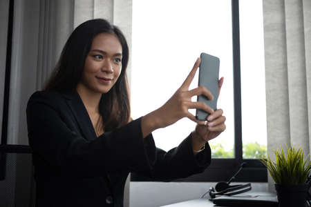 Portrait of charming businesswoman sitting near window in office and using mobile phone.