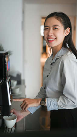 Portrait of happy young woman employee smiling and making coffee with coffee machine during break in office.の写真素材