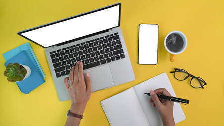 Above view of woman creative designer using laptop computer and making note on notebook over yellow...