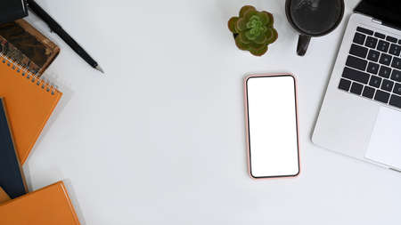 Top view workspace with empty screen of mobile phone and office supplies on white office desk.