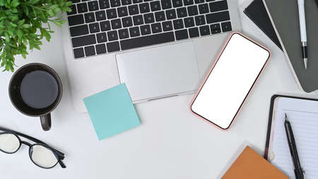 Stylish workspace with smart phone, laptop, notebook, glasses and flowers on orange background.
