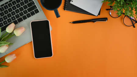 Stylish workspace with smart phone, laptop, notebook, glasses and flowers on orange background.の写真素材
