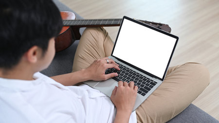 Young asian man using laptop computer while sitting with his guitar on couch at home.の写真素材