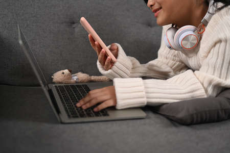 Cropped shot of happy young woman using smart phone and laptop while lying on sofa.の写真素材
