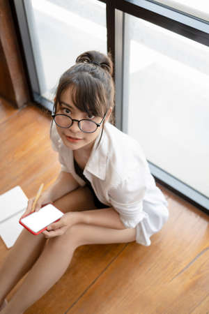 Young asian woman in eyeglasses holding mobile phone and looking at camera while sitting on wooden...