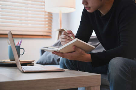 Cropped shot of young man using laptop computer and making note on notebook while sitting on sofa.