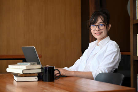 Cheerful female student sitting in library with laptop computer and smiling to camera.の写真素材
