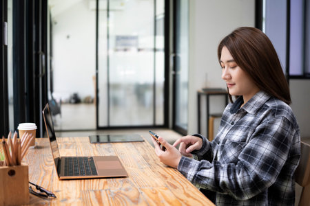 Side view young woman sitting in front of laptop at office desk and using mobile phone.の写真素材