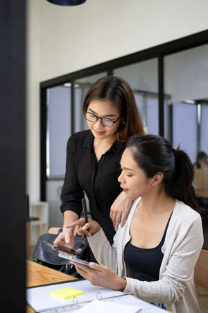 Two female colleagues sharing idea and using smart phone together in modern office.の写真素材