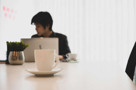 A white cup of coffee on white office desk with businessman sitting in background.の写真素材
